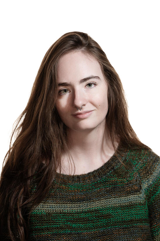 A headshot of Lucy Doherty, a young white woman in a green sweater with long brown hair and a septum piercing.